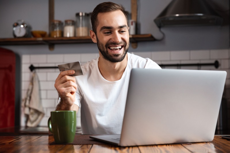 un jeune homme souriant, assis à un bureau profite de son cadeau homme sous la forme d'une carte cadeau pour effectuer du shopping en ligne