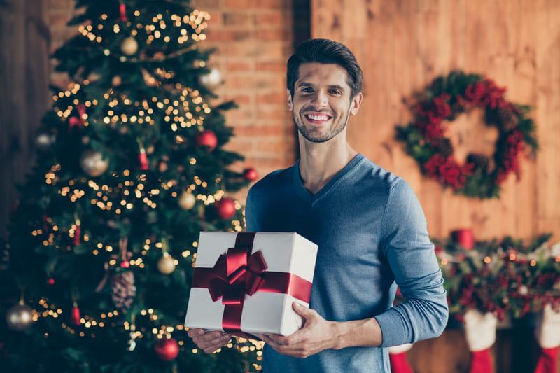 un homme qui aime le rugby se tient debout avec un cadeau entre les mains, il y a un sapin de noel et des décorations de noel en arriere plan 