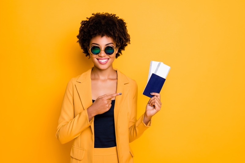 jeune femme aux cheveux afro sur fond jaune tient en main son passeport et ses billets d'avion pour son départ en vacances